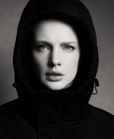 Monochrome portrait of a fair-skinned adult female with a hooded coat, face forward, with neutral expression against a grey background.