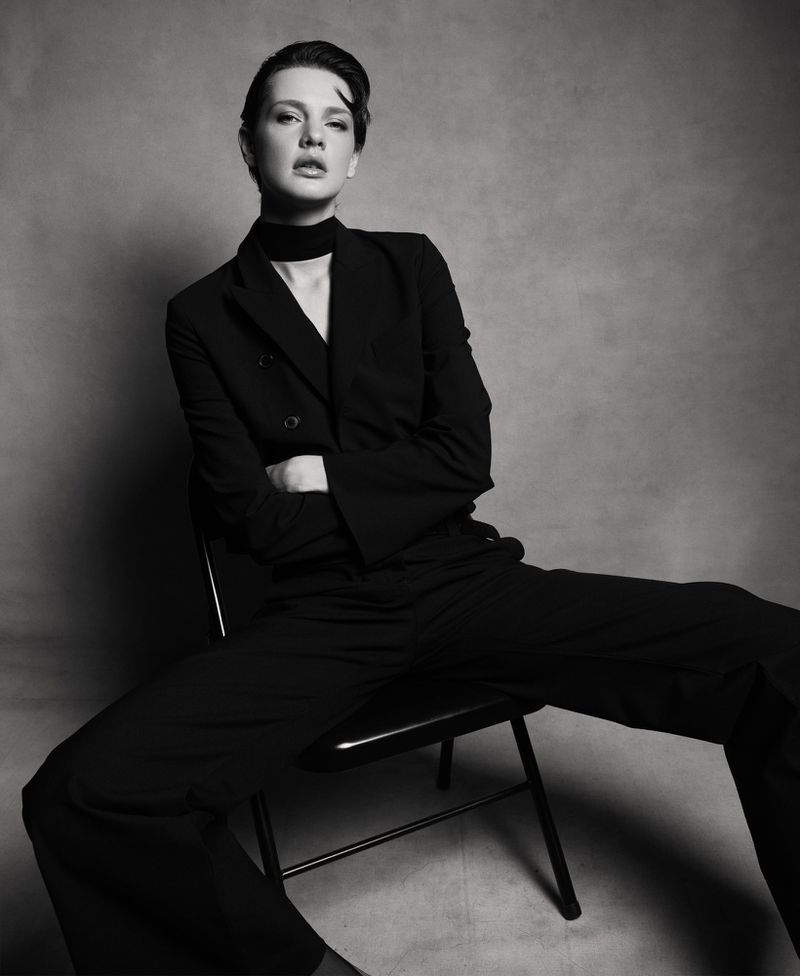 Black and white portrait of a woman in a suit sitting on a chair with arms crossed, facing forward in a photo studio setting.
