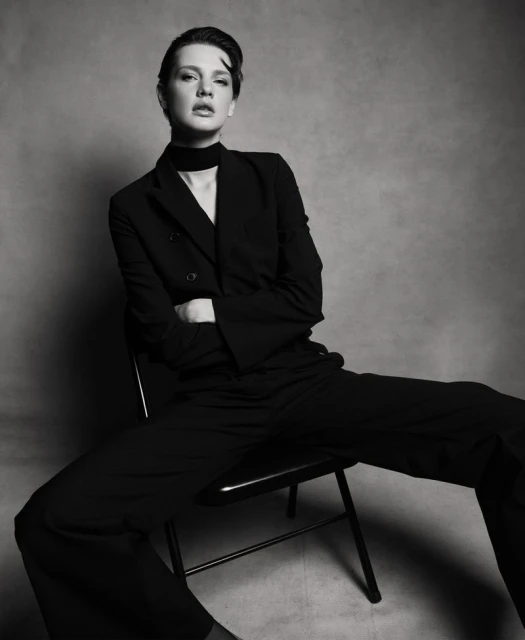 Black and white portrait of a woman in a suit sitting on a chair with arms crossed, facing forward in a photo studio setting.
