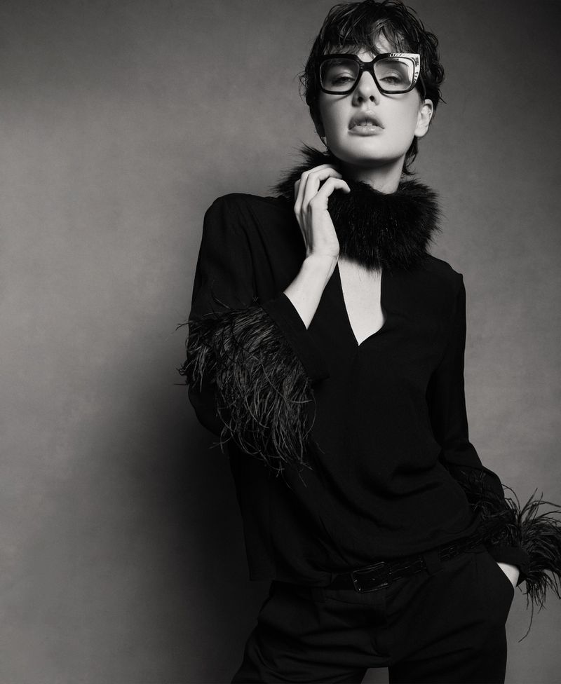Monochrome fashion shot of a stylish woman with short hair, wearing glasses and a feathered top, striking a confident pose indoors.