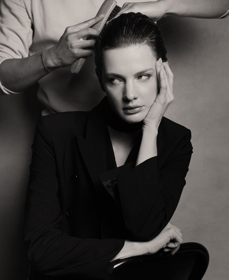 A young woman in a black blazer sits as someone brushes her hair back in a black and white photo studio setting with a grey background.
