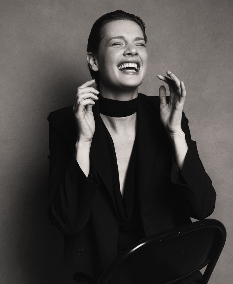 Black and white portrait of a laughing adult woman wearing a blazer and choker, sitting on a chair in a photo studio.