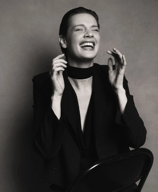 Black and white portrait of a laughing adult woman wearing a blazer and choker, sitting on a chair in a photo studio.