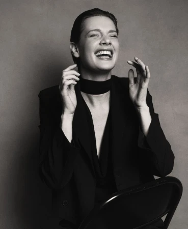 Black and white portrait of a laughing adult woman wearing a blazer and choker, sitting on a chair in a photo studio.