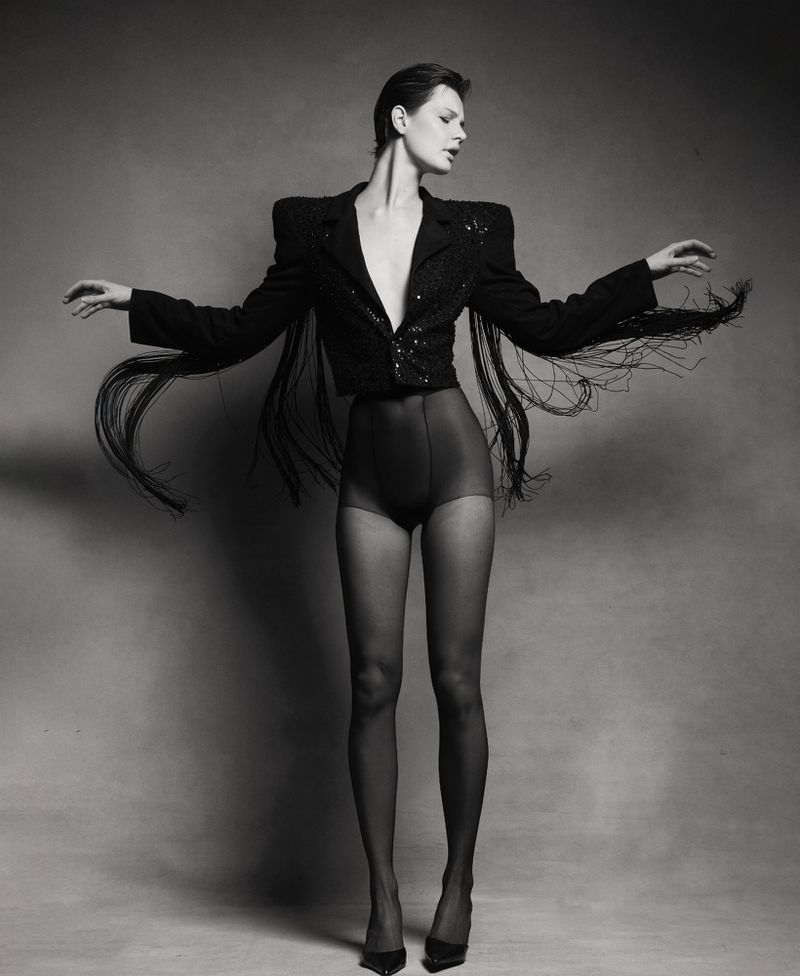 Monochrome full studio shot of a woman in a fringe jacket, dark shorts, and hosiery with arms outstretched against a grey background.