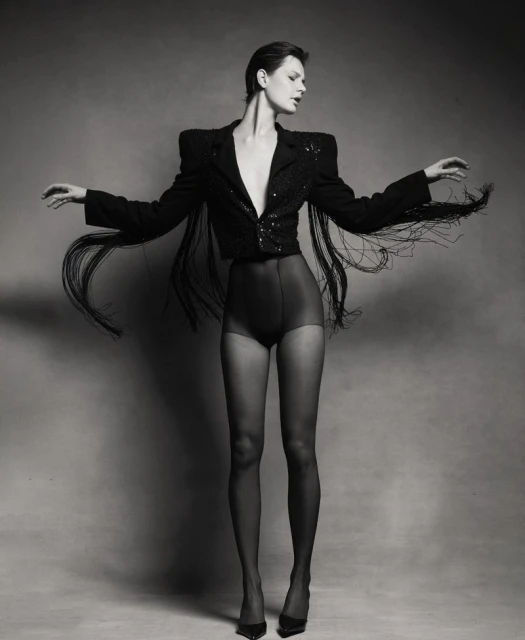 Monochrome full studio shot of a woman in a fringe jacket, dark shorts, and hosiery with arms outstretched against a grey background.