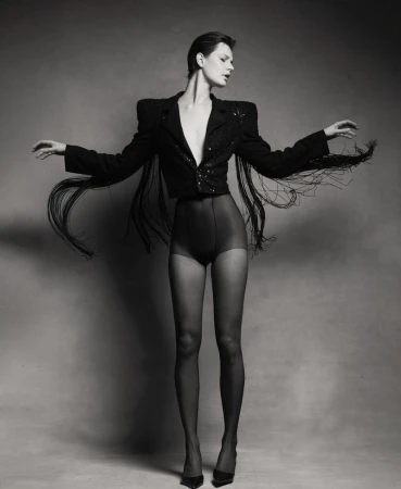 Monochrome full studio shot of a woman in a fringe jacket, dark shorts, and hosiery with arms outstretched against a grey background.