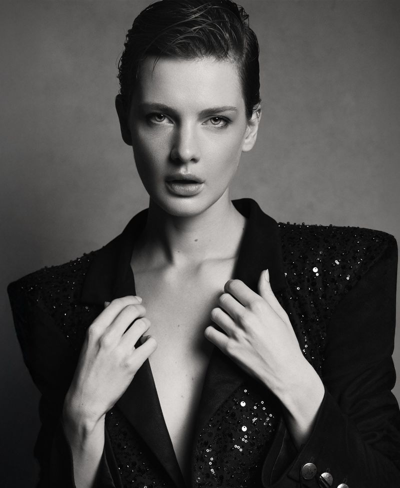 Monochrome portrait of a woman with short hair in a sequined blazer, hands at her collar, set against a grey studio backdrop.