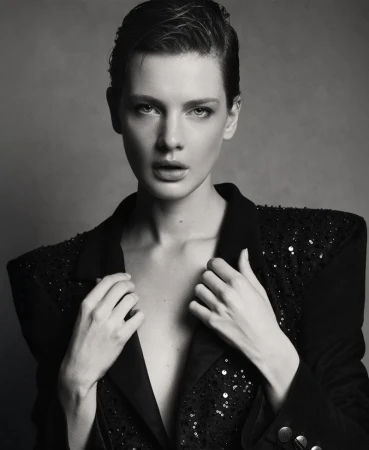 Monochrome portrait of a woman with short hair in a sequined blazer, hands at her collar, set against a grey studio backdrop.