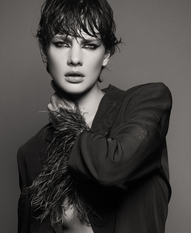 Black-and-white portrait of a female model with short, wet hair, dramatic eye makeup, and a feathered collar, posing in a photo studio.