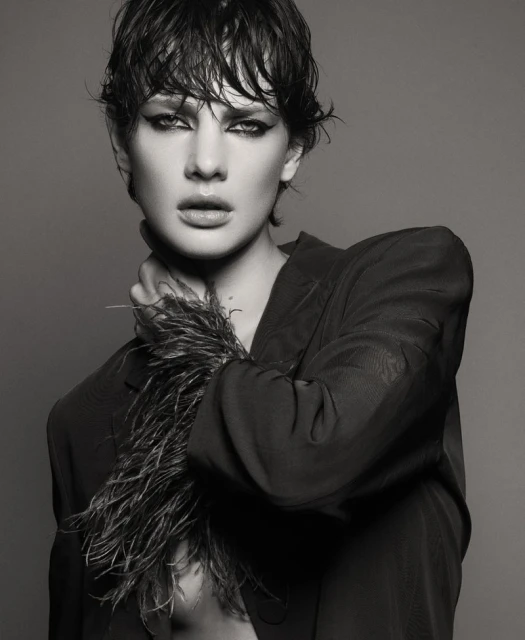 Black-and-white portrait of a female model with short, wet hair, dramatic eye makeup, and a feathered collar, posing in a photo studio.