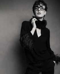 Model Mirthe Dijkstra wearing black rectangular eyeglasses and a feathered black blouse against a neutral studio backdrop.