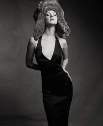 Monochrome portrait of a woman wearing an oversized textured fur hat and a black plunge halter dress in a studio setting.
