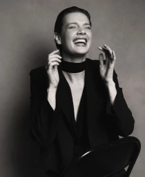 Mirthe Dijkstra laughing in a black tailored blazer and wide neck choker against a grey studio backdrop.