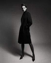 Model Mirthe Dijkstra posing in a structured black coat, sheer black tights, and pointed-toe heels against a grey studio backdrop.