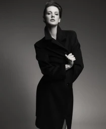 Monochrome studio portrait of model Mirthe Dijkstra wearing a structured black overcoat against a smooth grey background.