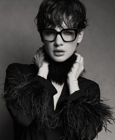 Monochrome portrait of model Mirthe Dijkstra wearing thick black eyeglasses and a feather-trimmed black top.