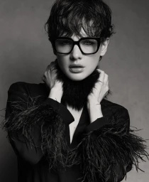 Monochrome portrait of model Mirthe Dijkstra wearing thick black eyeglasses and a feather-trimmed black top.