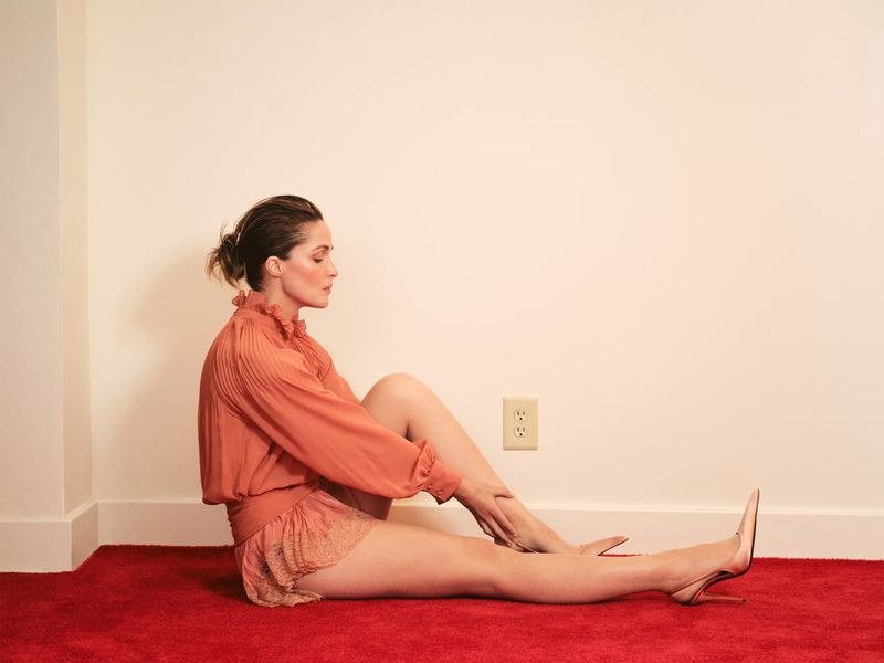 Woman with hair in updo sits on a red carpet in front of a cream wall wearing a peach-colored top and high-heeled shoes.