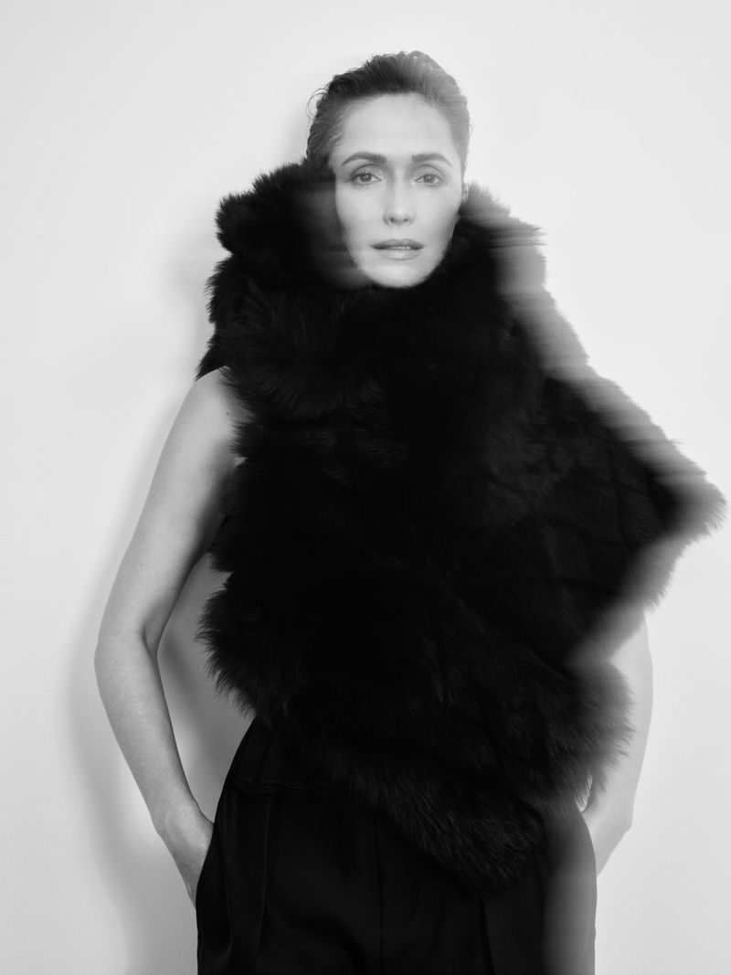 Black-and-white portrait of a woman in a dark outfit with fur shawl posing against a white backdrop with soft, diffused lighting.