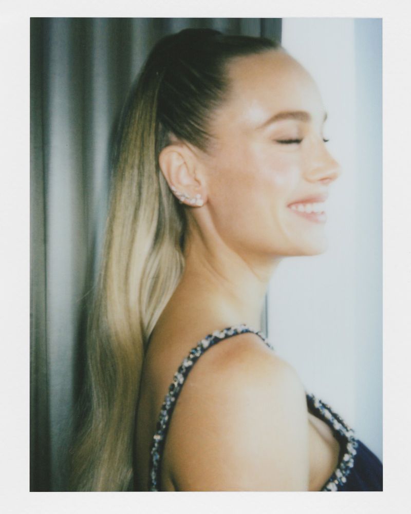 Close-up studio shot of a smiling adult woman with blonde hair in a high ponytail, wearing a sleeveless beaded top.