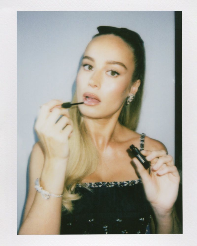 Close-up portrait of a fair-skinned woman applying lip gloss in a photo studio, wearing a black top, against a light blue background.
