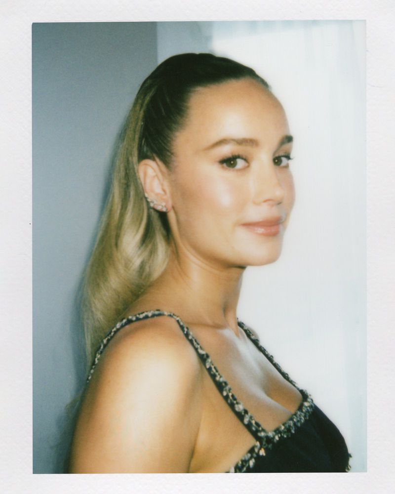 Portrait of a blonde woman with her hair in a ponytail, wearing a black dress with beaded straps, posing against a gray wall.