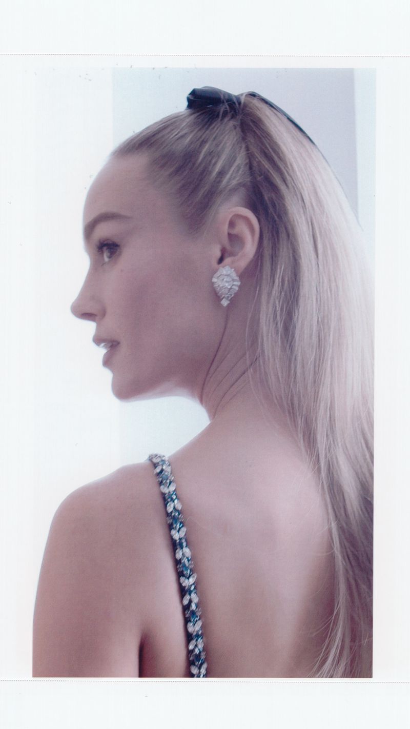 Blonde woman with a high ponytail and floral-strapped dress looks to the side against a bright, neutral background, wearing teardrop earrings.