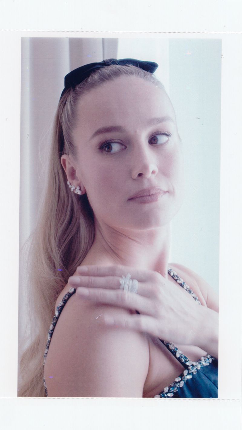 Blonde woman with a bow in her hair, wearing earrings and a ring, poses in a shoulder-up shot in front of white walls.