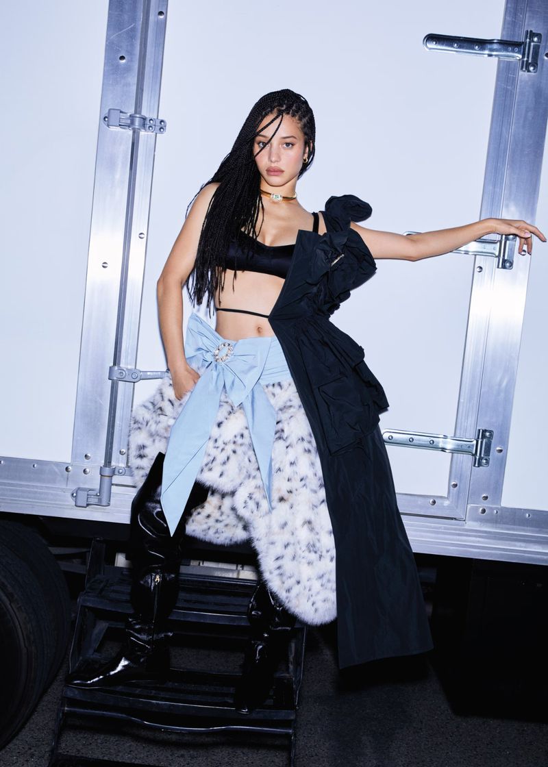 Fashion shot of a woman in a black top, light blue skirt with a bow, and thigh-high black patent boots, standing on metal stairs.