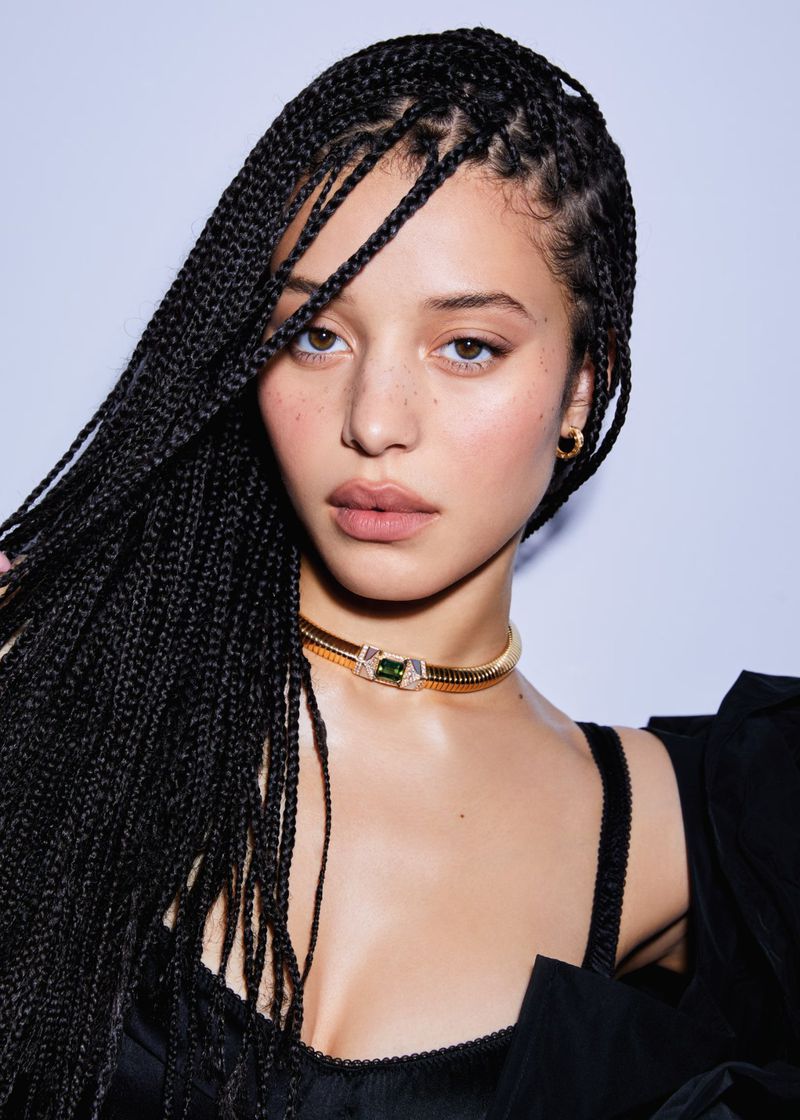 Portrait of a young adult female with box braids, wearing a gold choker, against a light gray studio backdrop, shot with soft lighting.