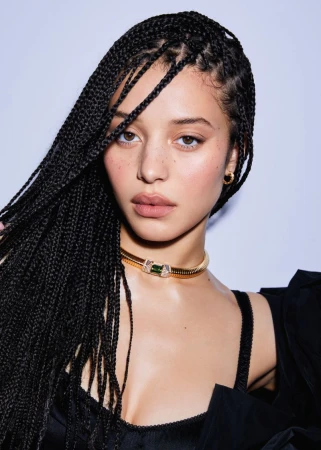 Portrait of a young adult female with box braids, wearing a gold choker, against a light gray studio backdrop, shot with soft lighting.