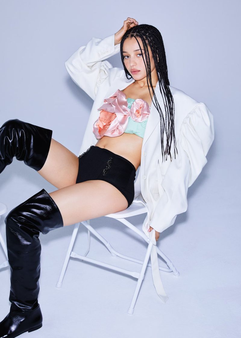 A woman with braided hair sits on a white folding chair wearing a white blazer, floral crop top, black shorts, and high black boots.