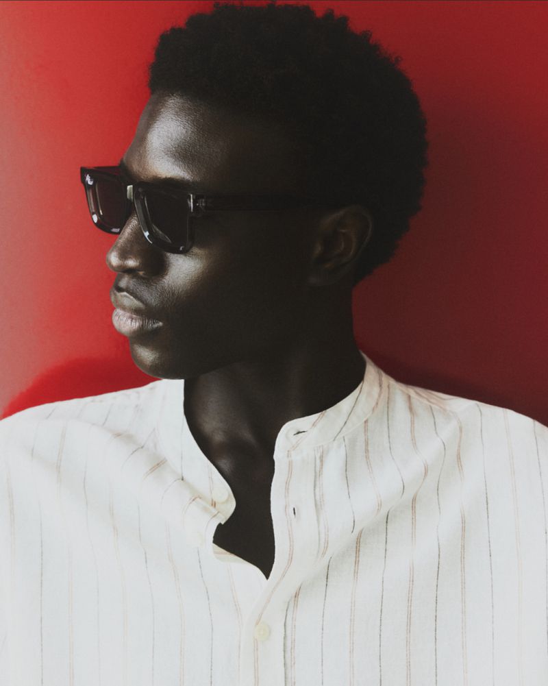 Profile shot of a dark-skinned male wearing a white button-down shirt and sunglasses against a red background in a photo studio.