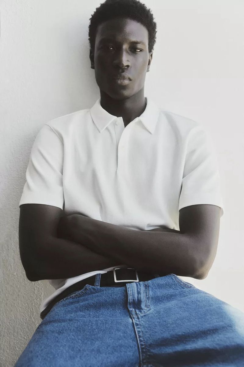 Black man with crossed arms wearing a white polo shirt and blue jeans against a plain wall