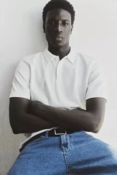Black man with crossed arms wearing a white polo shirt and blue jeans against a plain wall