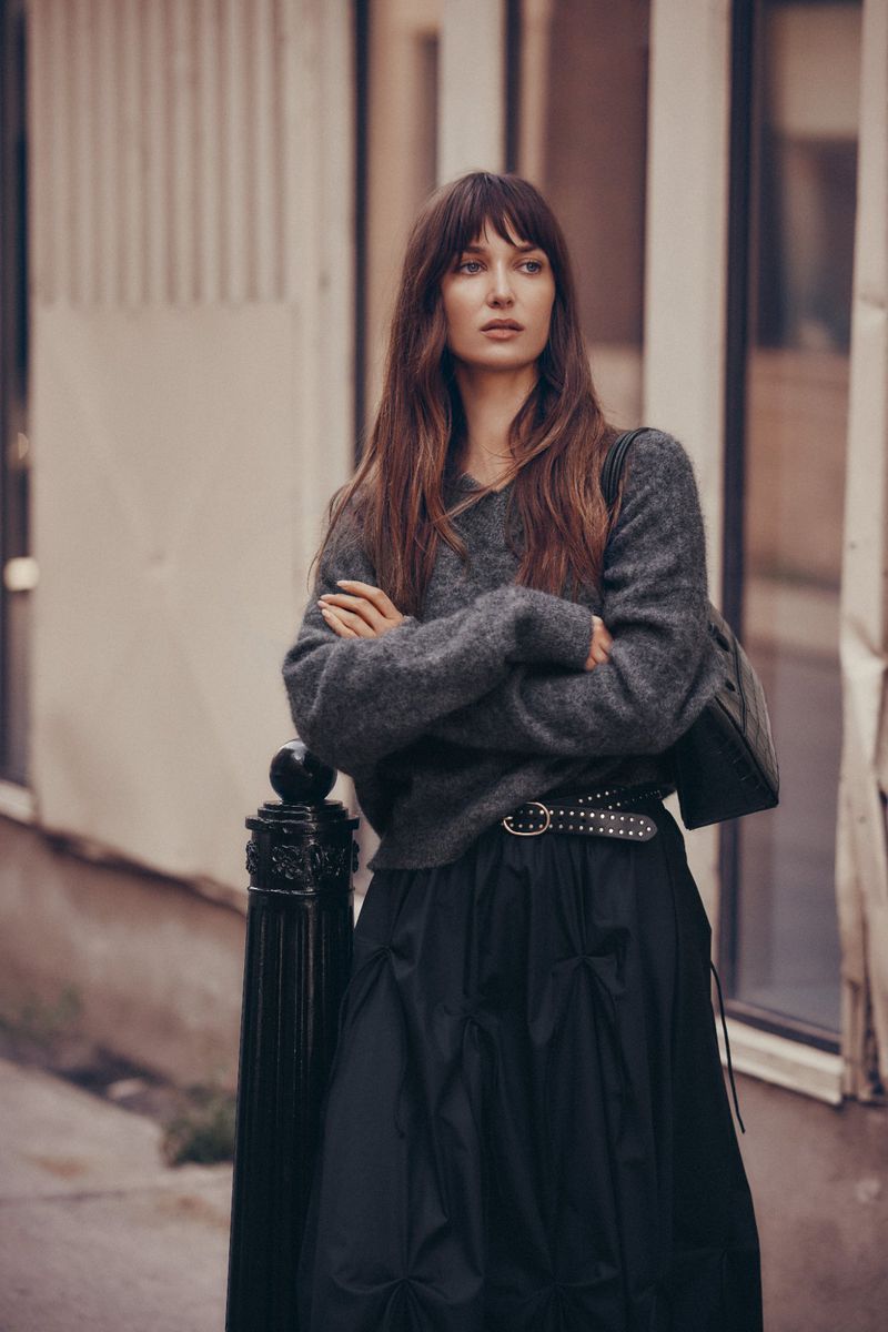 A woman with long brown hair stands with arms crossed in an urban setting, wearing a grey sweater and a black skirt with a studded belt.