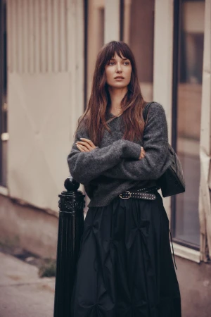A woman with long brown hair stands with arms crossed in an urban setting, wearing a grey sweater and a black skirt with a studded belt.