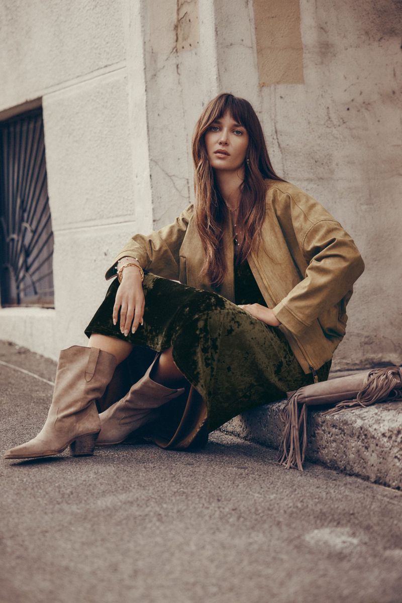 Adult woman in a tan jacket and olive green velvet dress and tan boots, sitting on the edge of a concrete step outdoors.