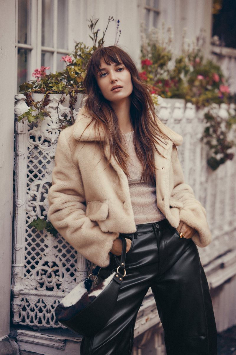 Model wearing a cream fluffy jacket and black trousers leaning against an ornate white fence.