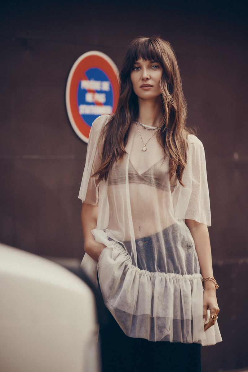 Brunette woman in sheer top poses against brown wall with a circular blue, white, and red sign in an eye-level outdoor shot.