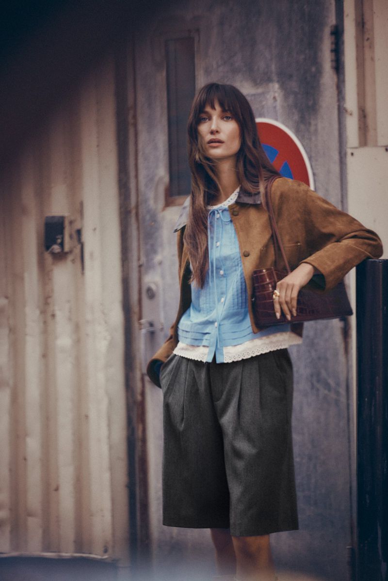 A stylish woman poses outdoors in a brown jacket, light blue top, and culottes, holding a clutch in front of a metal wall.