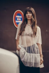 Model Ari Fournier wearing a sheer cream ruffled overlay top against an urban backdrop with a French parking sign.