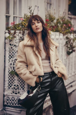 Model wearing a cream fluffy jacket and black trousers leaning against an ornate white fence.