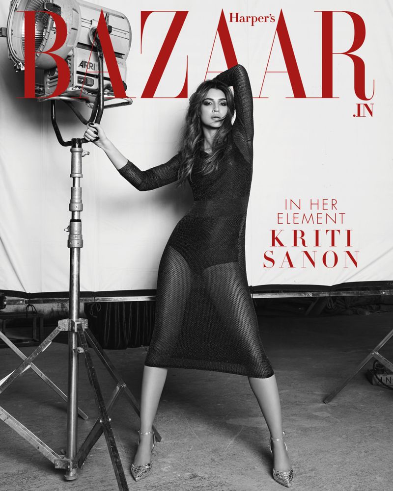 Black-and-white studio fashion shot of Kriti Sanon, wearing a sheer black dress and high heels, posing next to a large Arri light.
