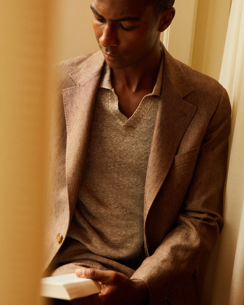Dark-skinned adult male in a brown suit and grey shirt indoors, looking down with soft lighting and neutral tones for fashion editorial.