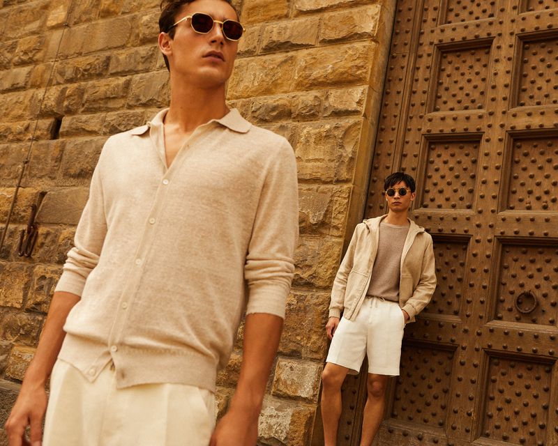 Two adult men pose against a stone wall and doorway wearing casual summer clothing in beige and white tones, stylish sunglasses.