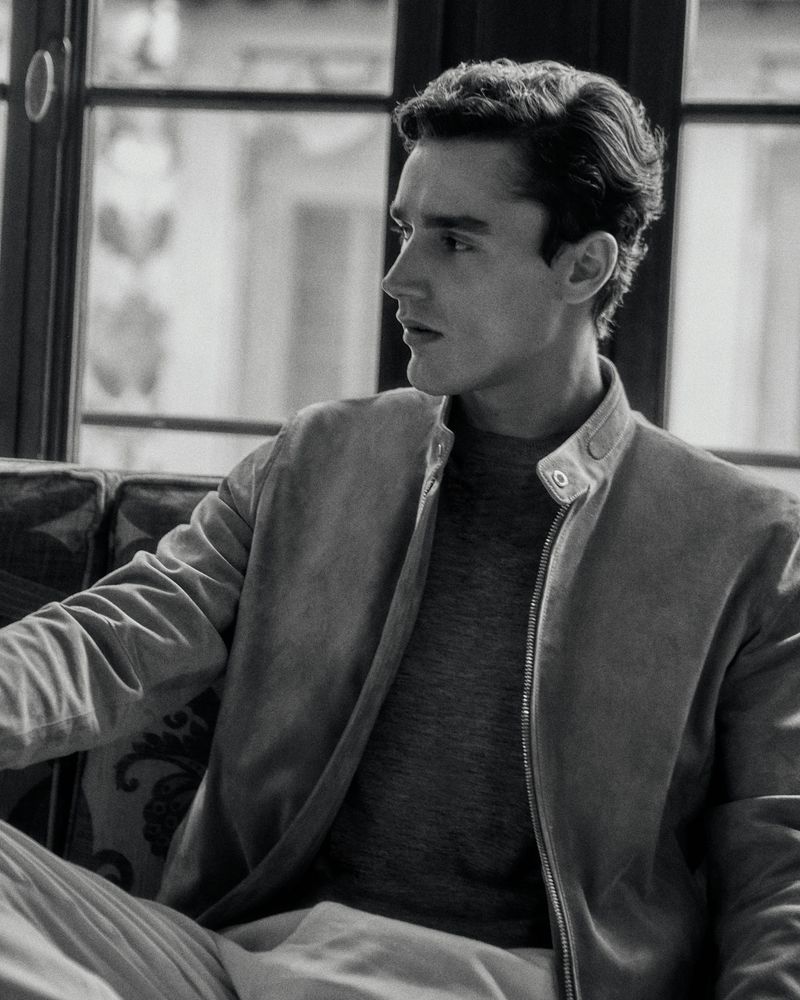 Black and white portrait of a young adult male indoors, wearing a casual jacket and sweater, sitting on a patterned couch near a window.