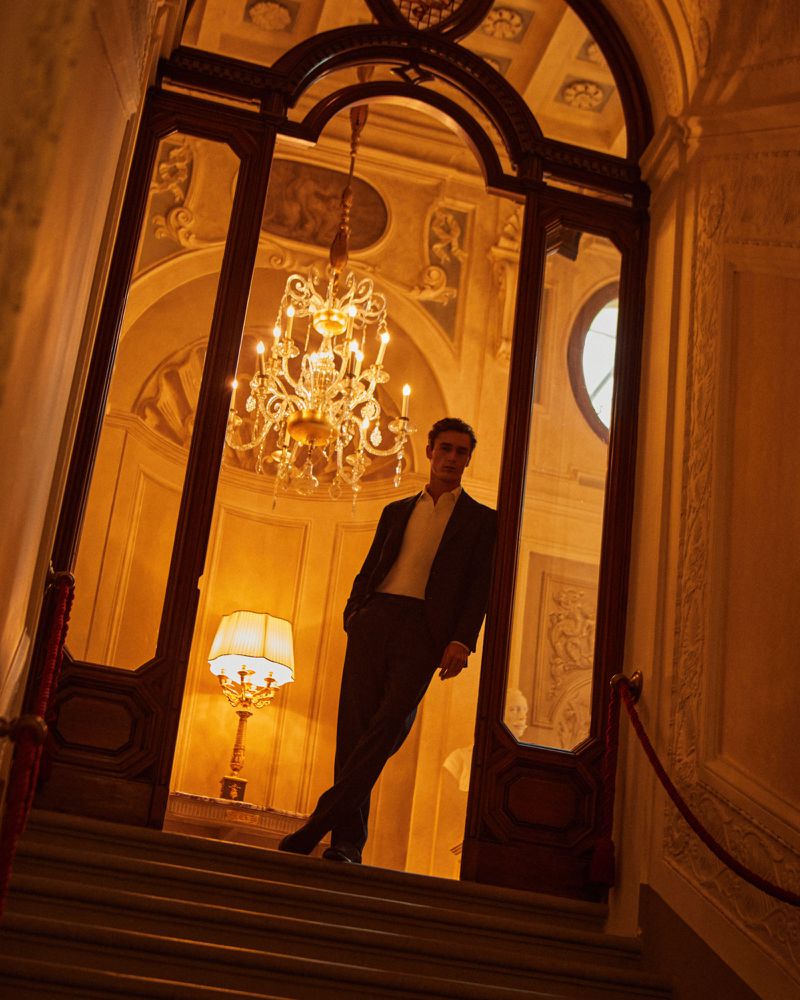 Man in suit stands in doorway of ornate building interior with chandelier and lamp, warm color grading, marble steps.