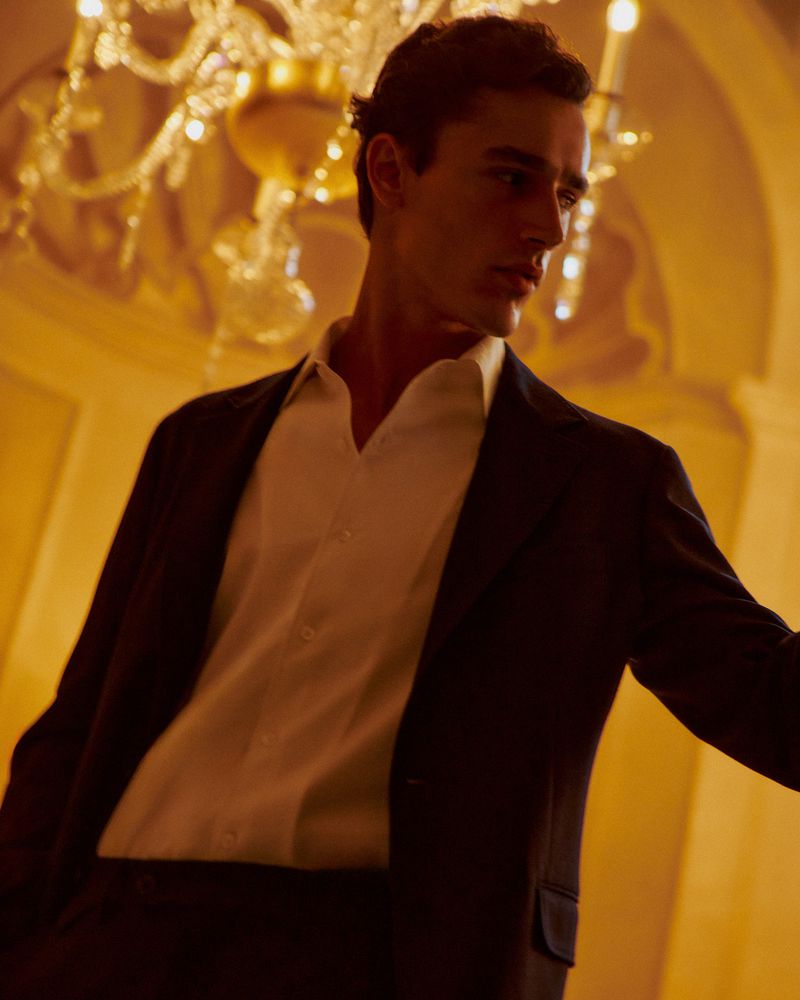 A well-dressed adult male poses indoors under a bright chandelier, wearing a suit jacket and a white collared shirt, with golden lighting.